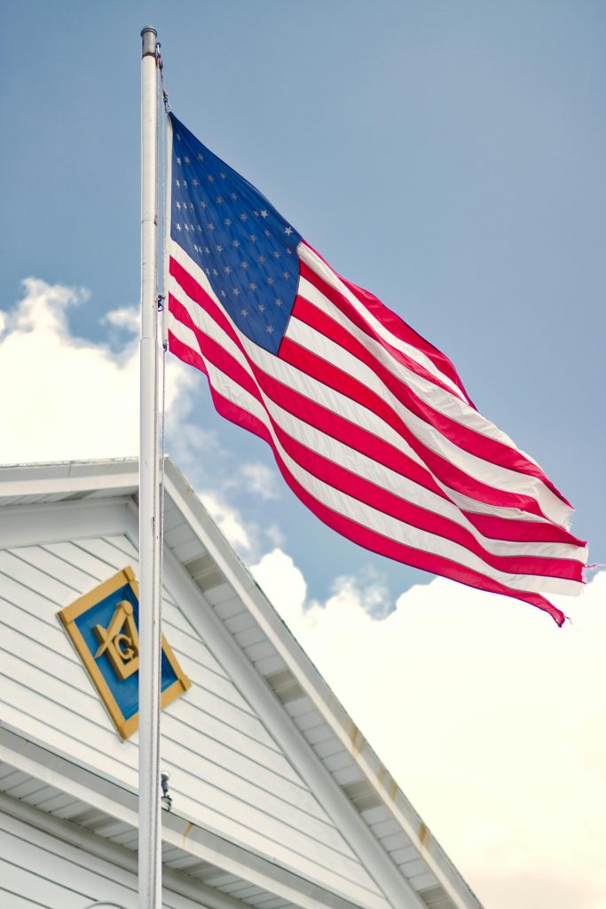 An American flag waves proudly in front of a Masonic Lodge against a clear blue sky.