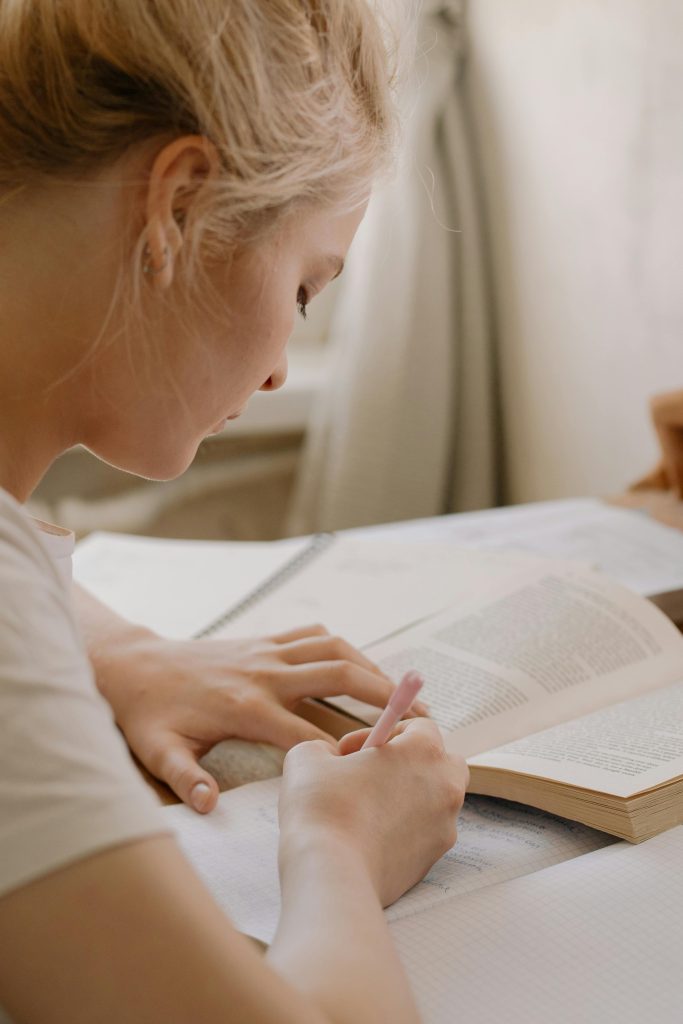 Focused young woman writing notes from an open textbook at a desk, creating a study environment.
